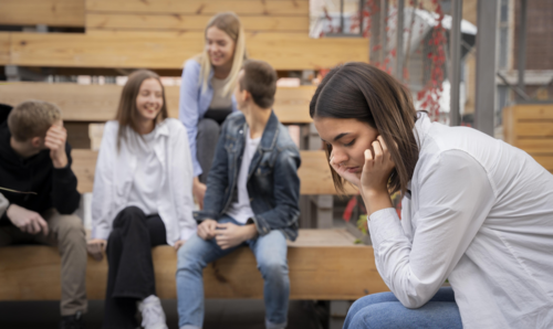 Eine Jugendliche wirkt belastet, während eine Gruppe Jugendlicher im Hintergrund lacht. Bild: Freepik