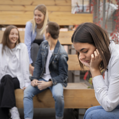 Eine Jugendliche wirkt belastet, während eine Gruppe Jugendlicher im Hintergrund lacht. Bild: Freepik